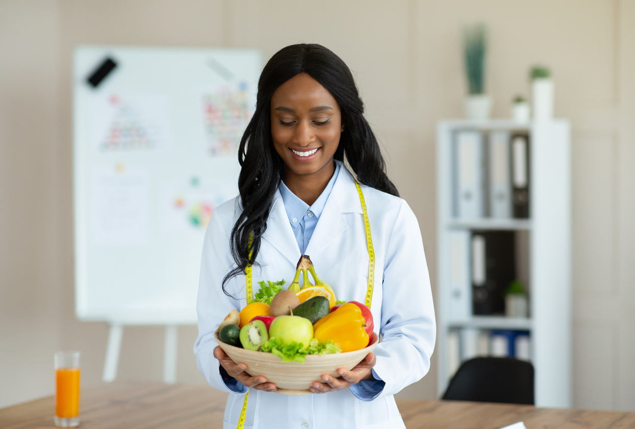 Portrait of joyful black nutritionist in lab coat holding bowl of fresh fruits and veggies at weight loss clinic. Healthy nutrition consultant recommending balanced plant based diet